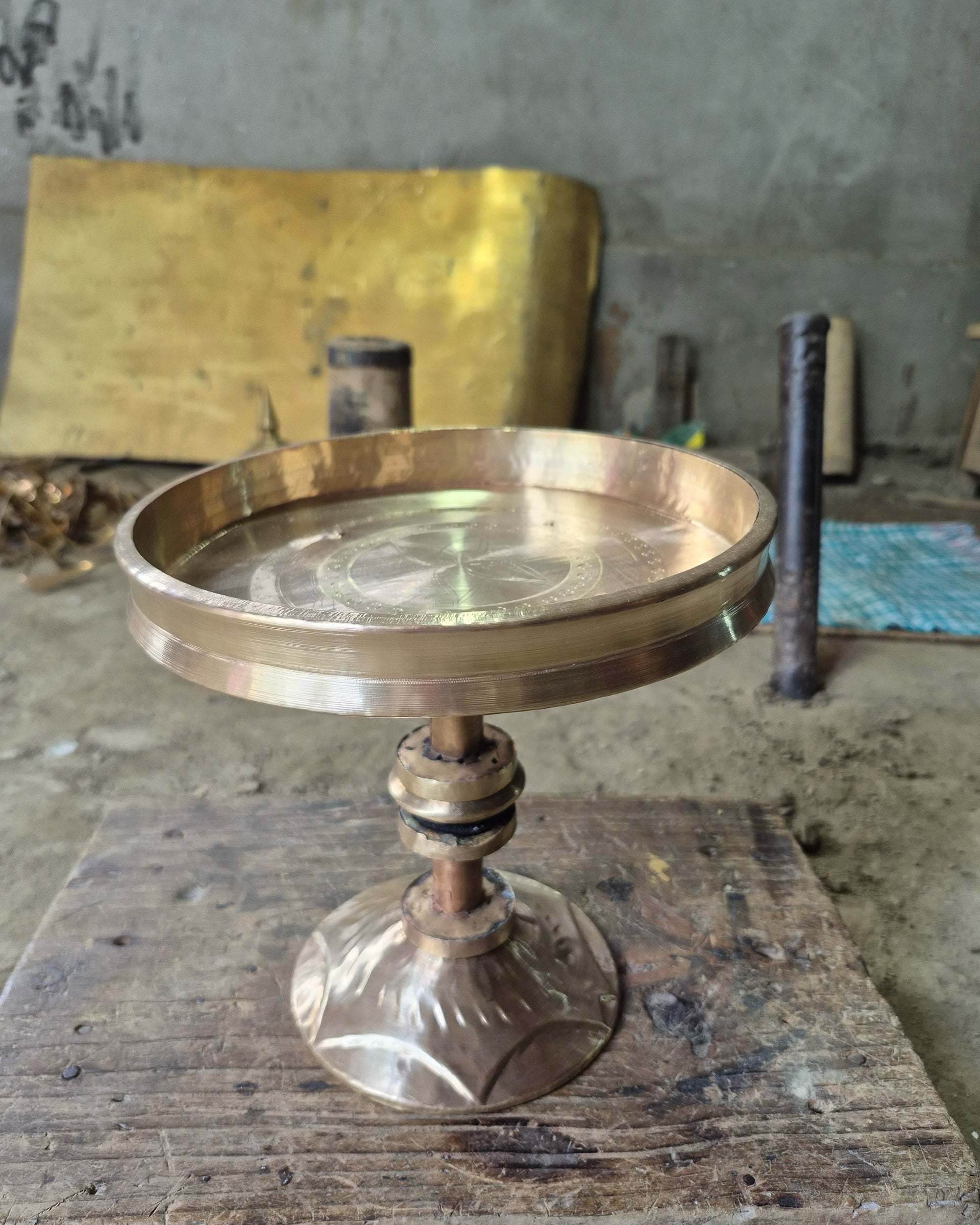 A lidless metal tray on a copper-coloured tube stem with collars and a lobed, flared foot, standing on a scarred wooden bench.