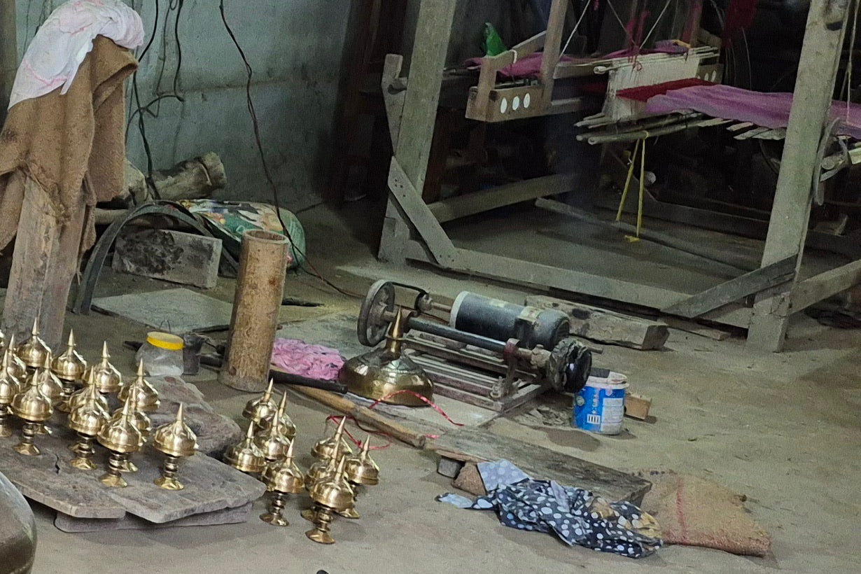 Rows of polished spire finials on boards, behind them a motor on a low frame beside a polished lid, and a handloom with pink cloth at the back.